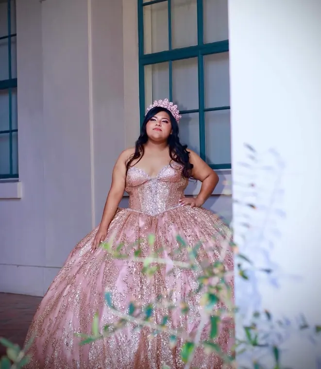 Quinceañera in a rose gold glitter gown posing by a green-trimmed window