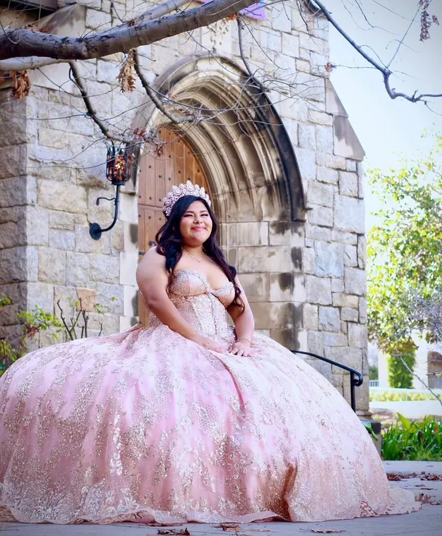 Quinceañera in a blush pink ball gown seated outside a stone chapel