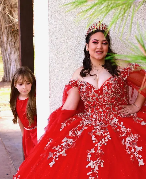 Quinceañera with a red crown leaning against a white wall